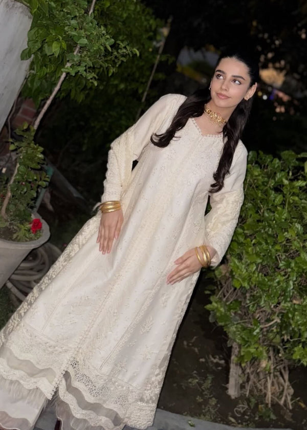 A woman wearing a long cream-colored embroidered kurta and matching skirt stands outdoors at night in front of green foliage. She is wearing gold jewelry and has her hair styled in a low bun.