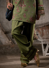 Person wearing a green traditional outfit with floral patterns and gold shoes in an urban setting.
