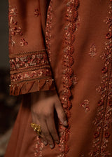Close-up of a hand wearing a gold ring with an embroidered brown garment in the background