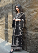 Woman in black embroidered traditional outfit standing against a stone wall.